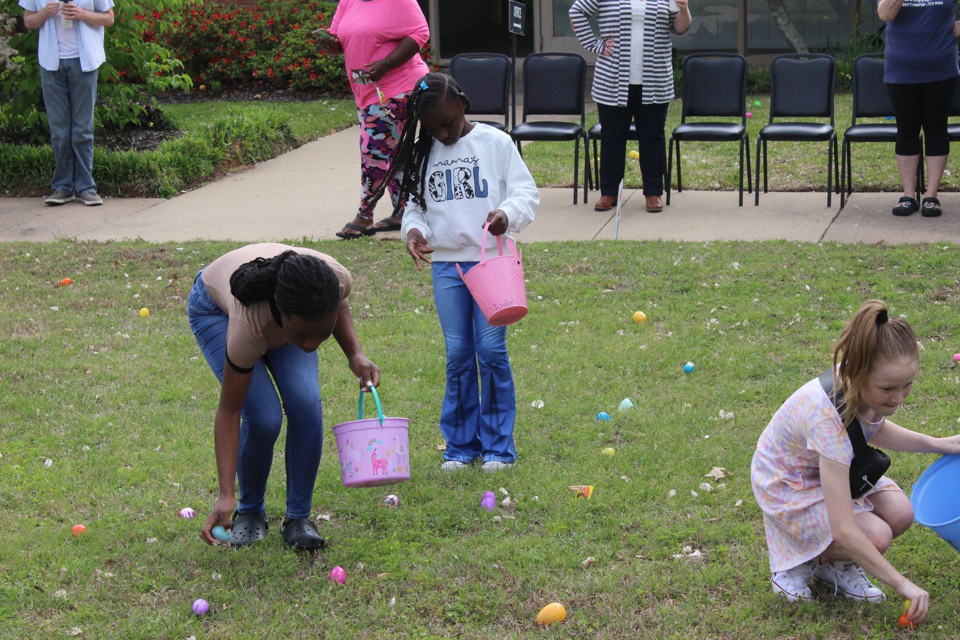 Children gathering Easter eggs on a lawn with adults standing nearby.