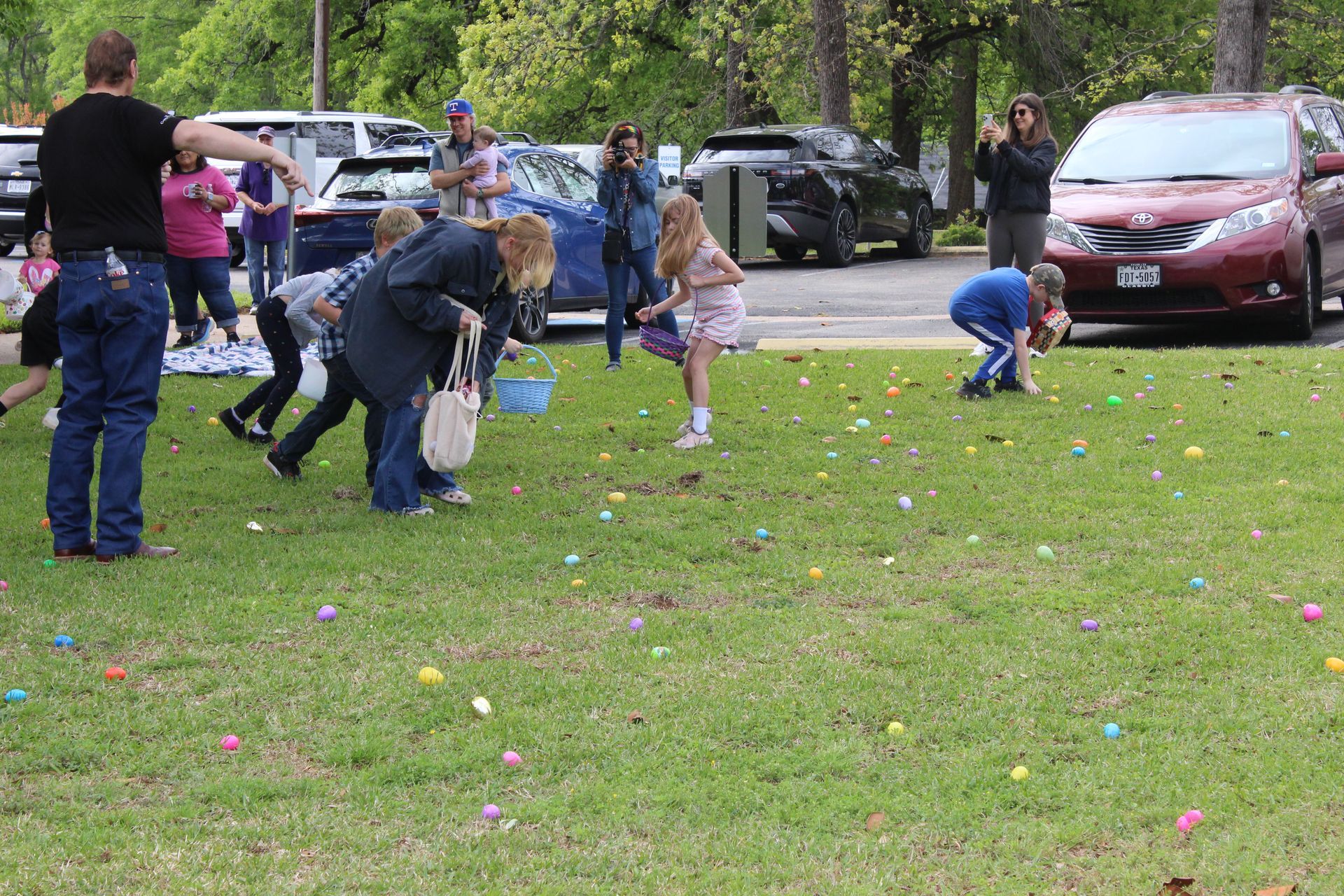 Children collecting Easter eggs on a grassy field; adults watch nearby.
