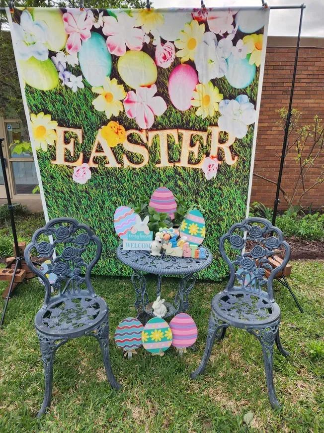 Easter-themed outdoor photo setup with backdrop of eggs and flowers, small table, chairs, and decorations.