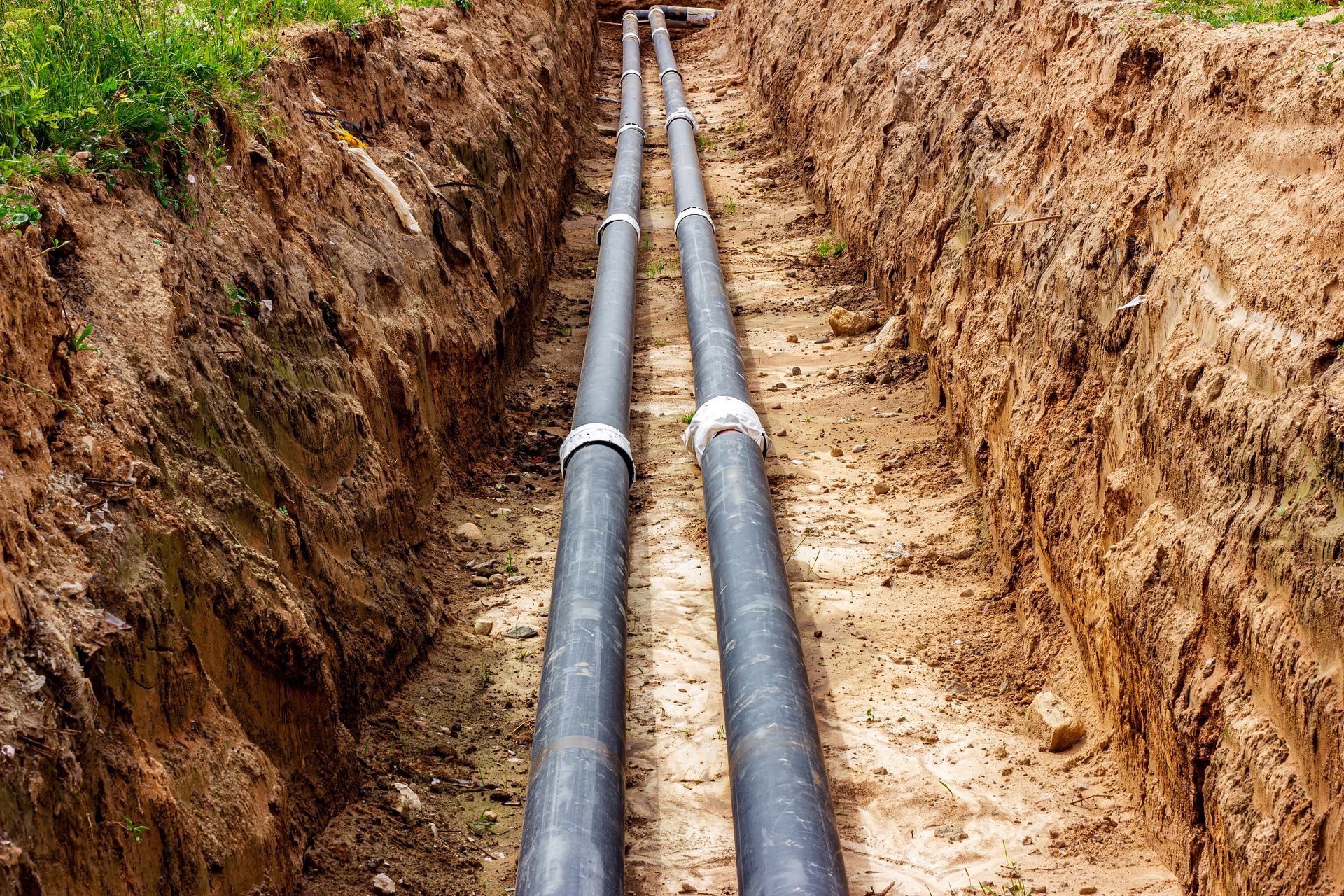 Two black pipes laid in a narrow, open trench, surrounded by brown soil and grass.