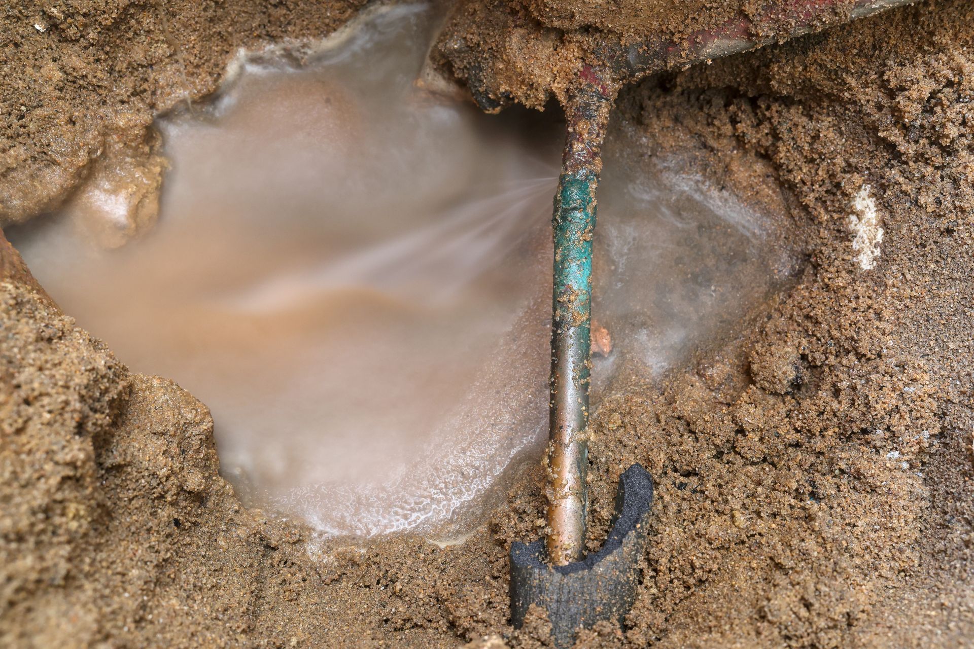 Water gushing from a corroded copper pipe buried in brown soil.
