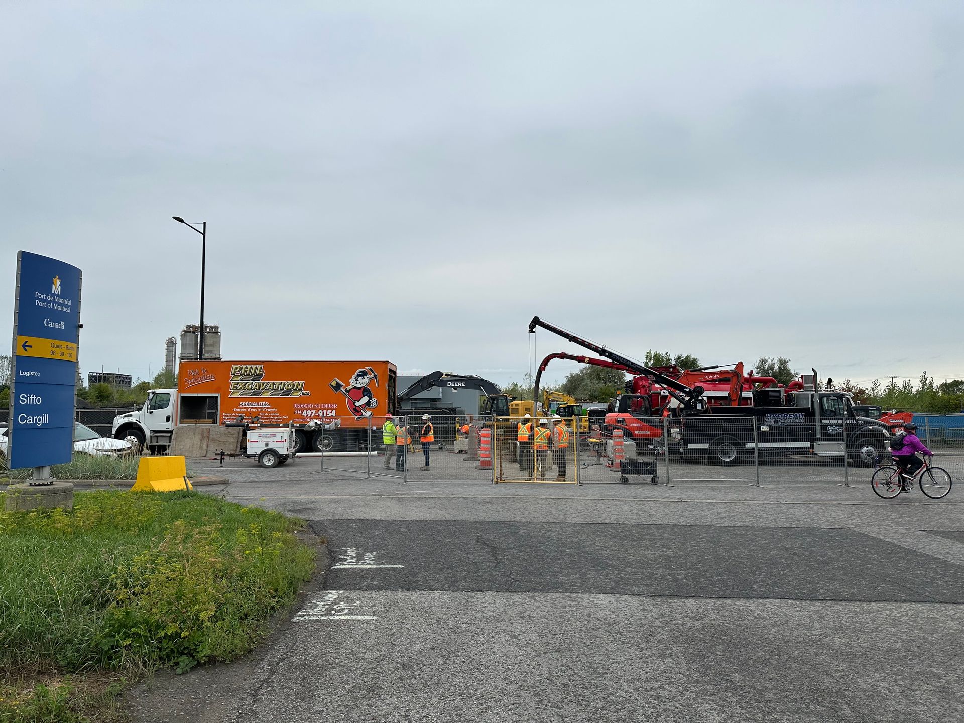 Construction site with trucks, workers, and a cyclist on a paved surface under an overcast sky.