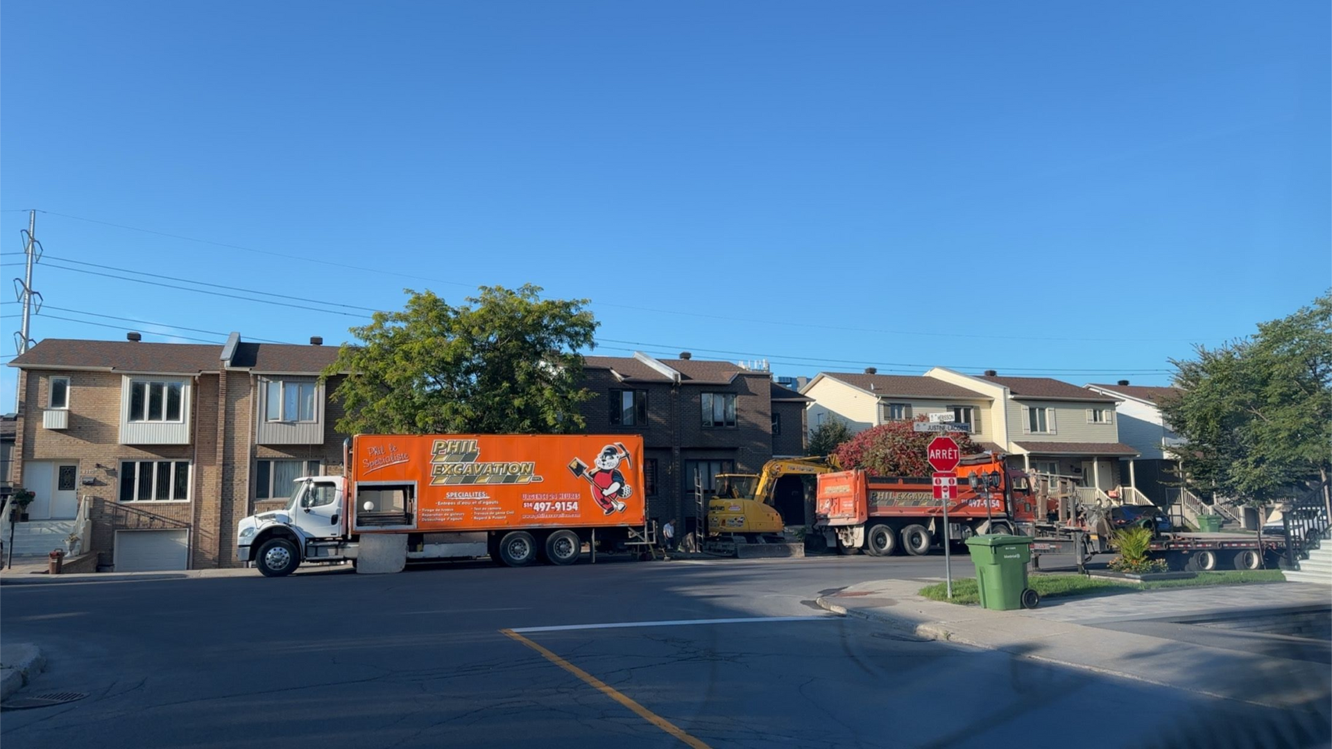 Des camions sont stationnés dans une rue bordée de maisons mitoyennes. L'un est orange, les autres sont jaunes et rouges. Ciel dégagé et ensoleillé.