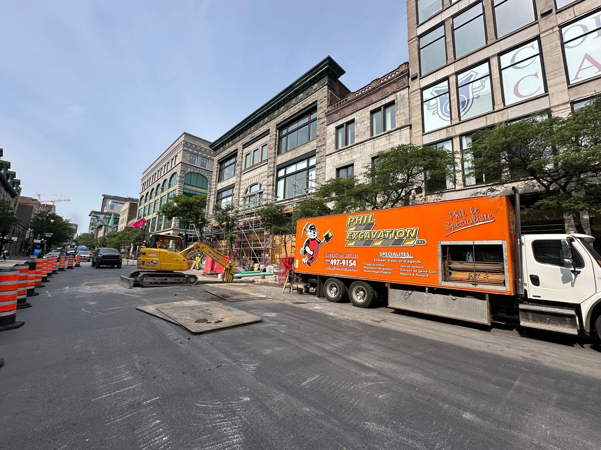 Street construction: Orange truck, excavator, and cones on a city street with multi-story buildings and shops.