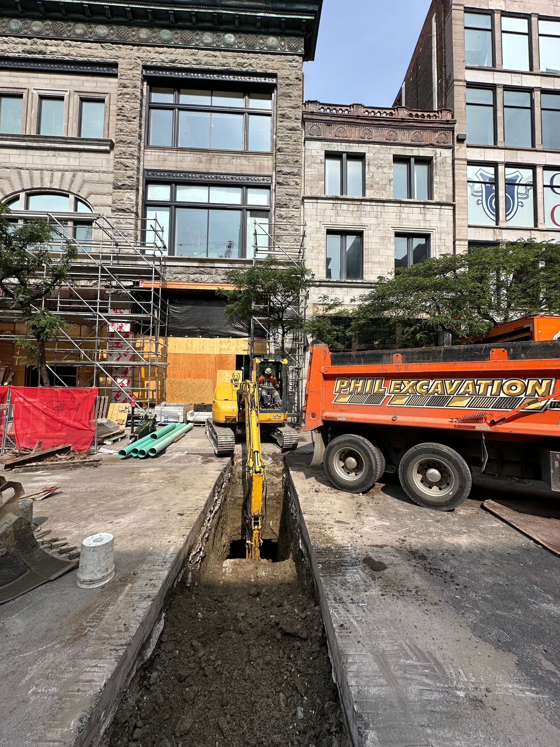 Chantier : Une pelleteuse creuse une tranchée dans une rue, un camion-benne se trouve à proximité. Des bâtiments en pierre sont visibles en arrière-plan.