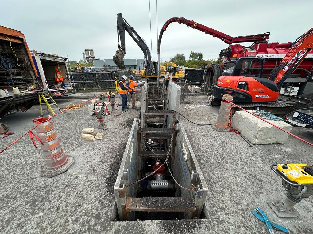 Chantier avec tranchée et engins de chantier. Présence d'ouvriers en gilets orange.