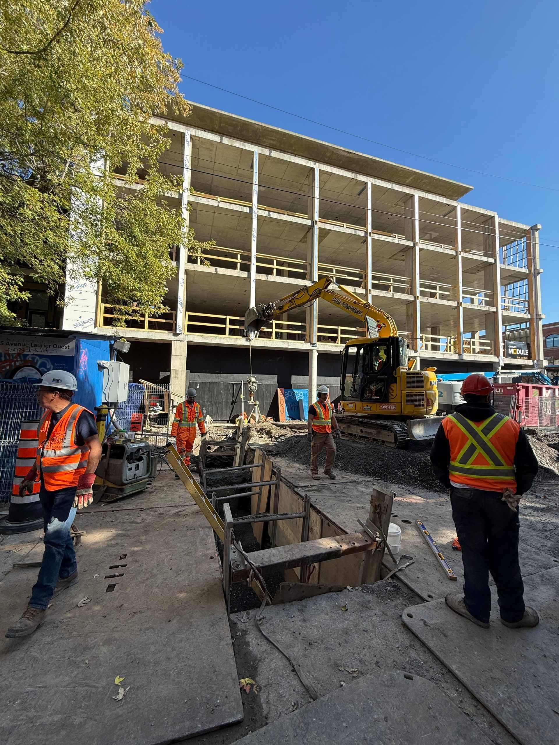 Construction site with workers, excavator, and partially built concrete building on a sunny day.