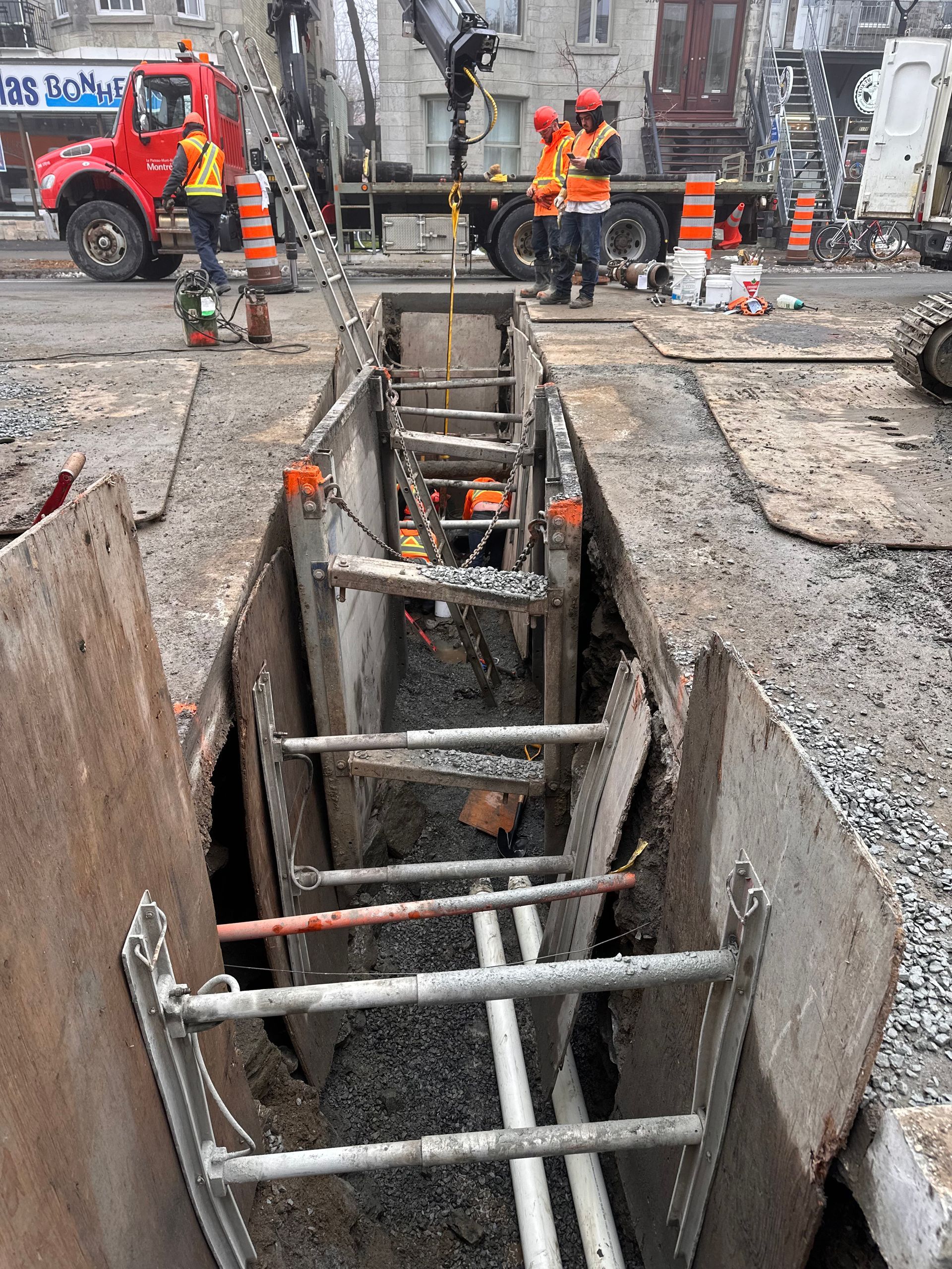 Construction site: Workers in orange vests beside a trench, equipment, and a red truck on a city street.