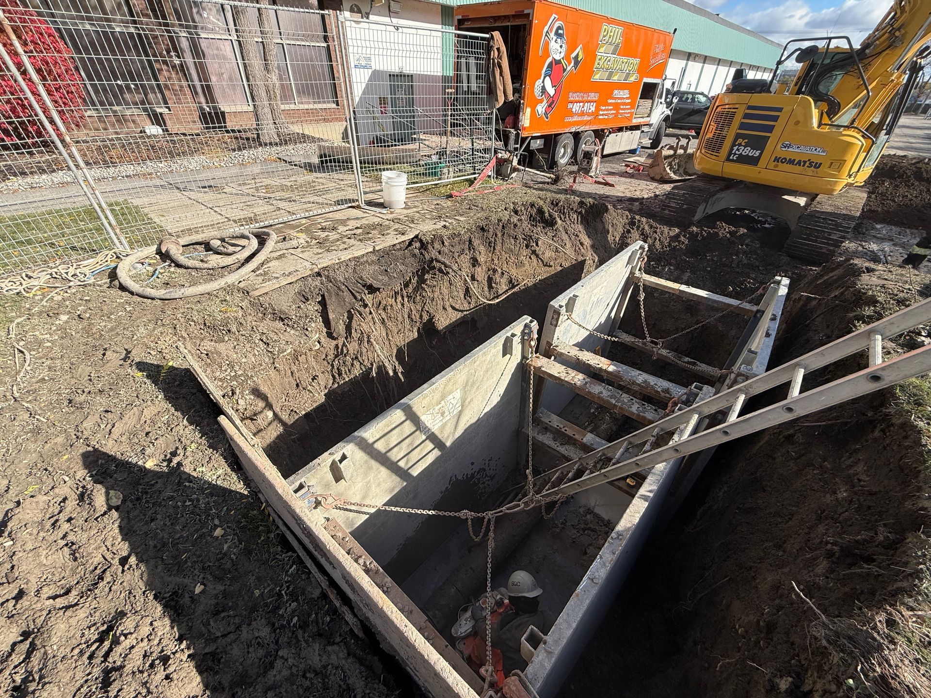 Chantier : tranchée avec étaiement, échelle et pelle mécanique. Un ouvrier travaille dans la tranchée. Un camion orange et un bâtiment sont visibles en arrière-plan.