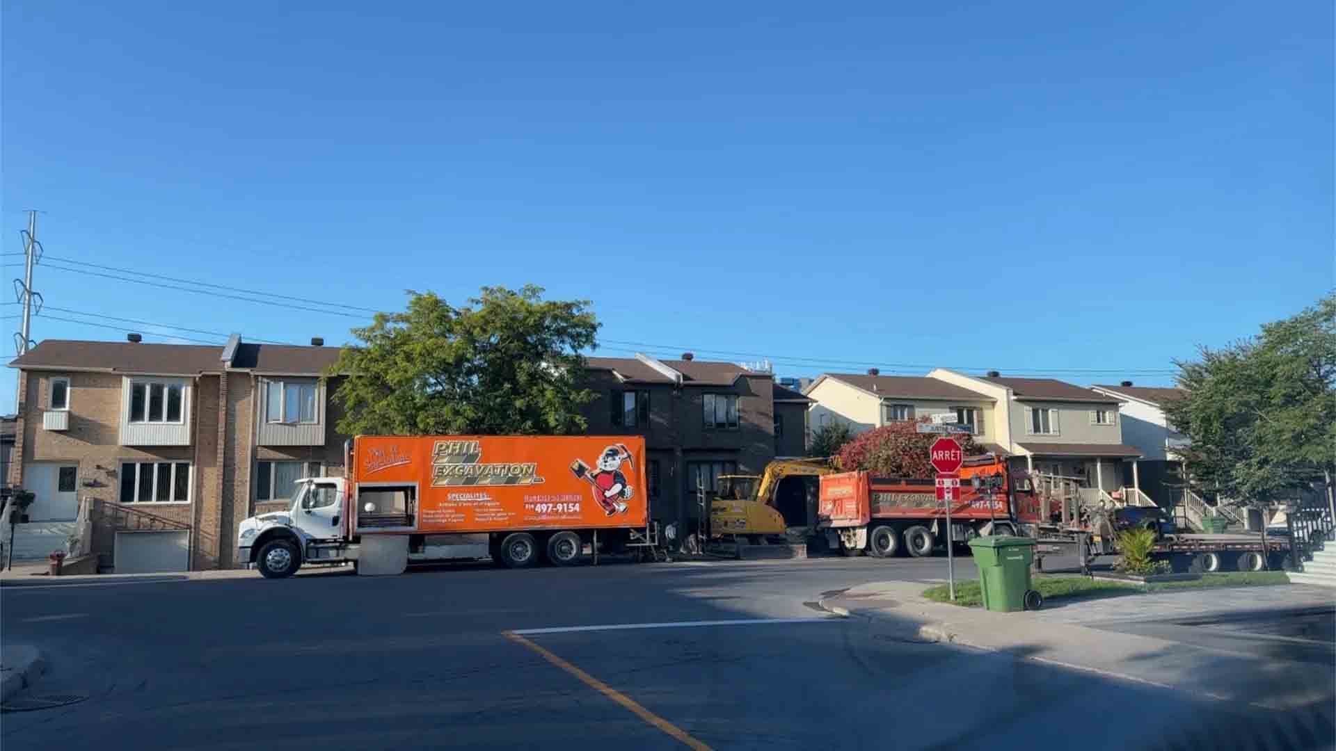 Des camions sont stationnés dans une rue bordée de maisons mitoyennes. L'un est orange, les autres sont jaunes et rouges. Ciel dégagé et ensoleillé.
