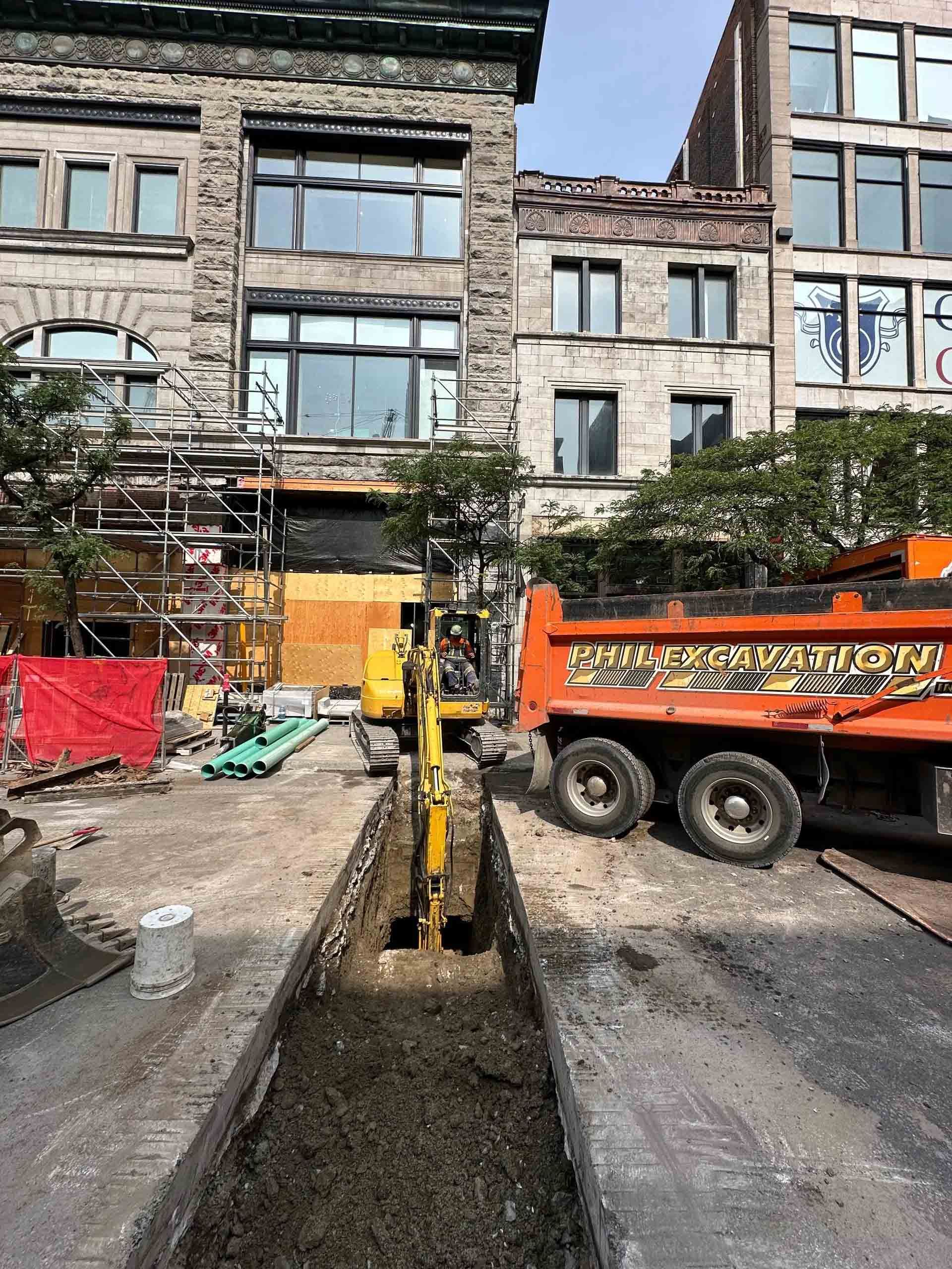 Chantier : Une pelleteuse creuse une tranchée dans une rue, un camion-benne se trouve à proximité. Des bâtiments en pierre sont visibles en arrière-plan.