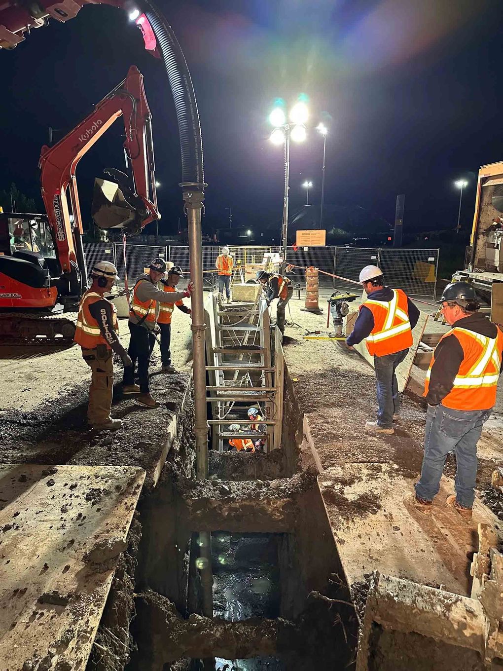 Des ouvriers du bâtiment travaillent de nuit près d'une profonde fosse d'excavation. Ils portent des gilets orange et des casques de chantier.