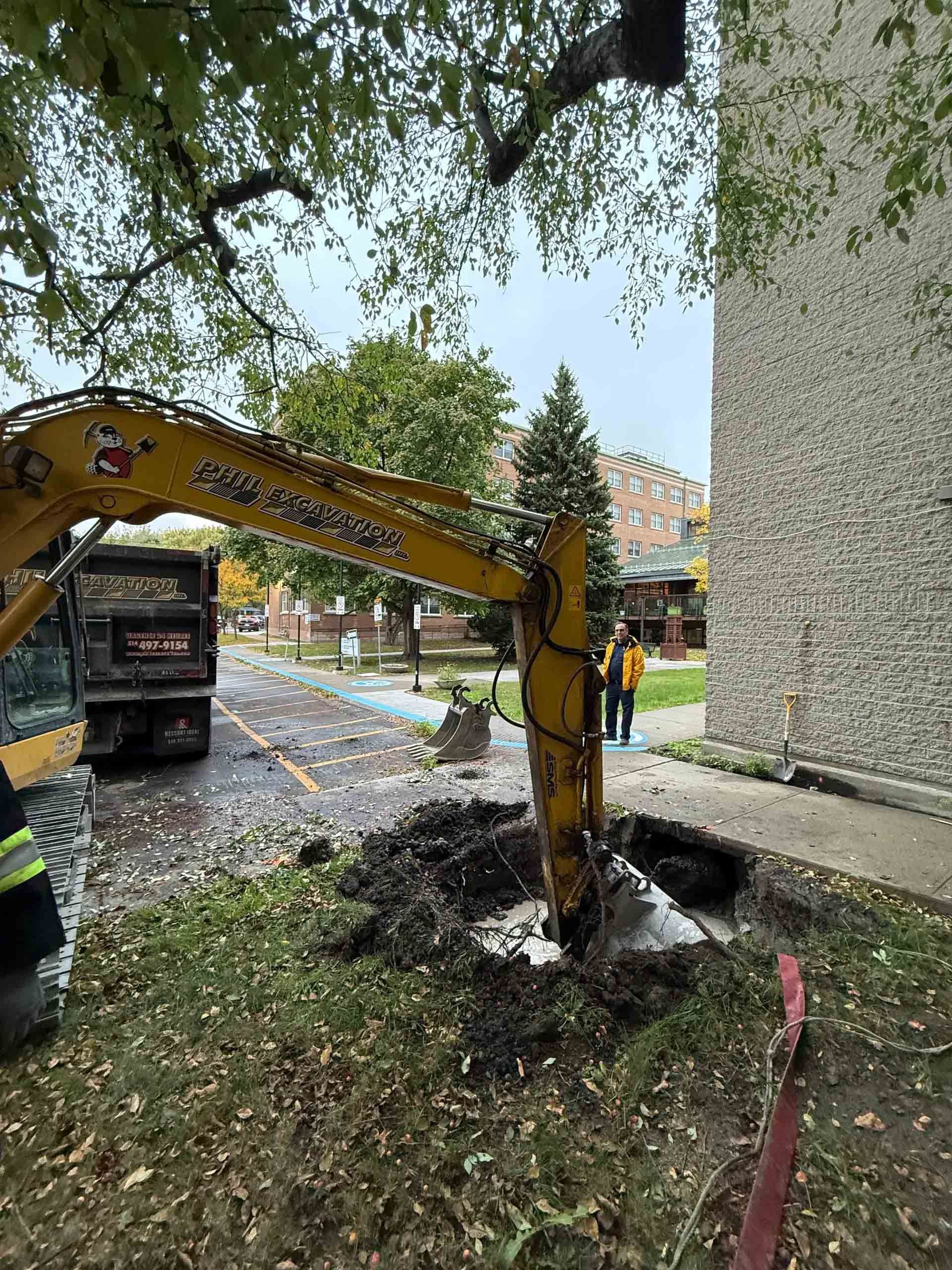 Une pelleteuse creuse près d'un bâtiment. Machine jaune, tas de terre, ouvriers qui regardent.
