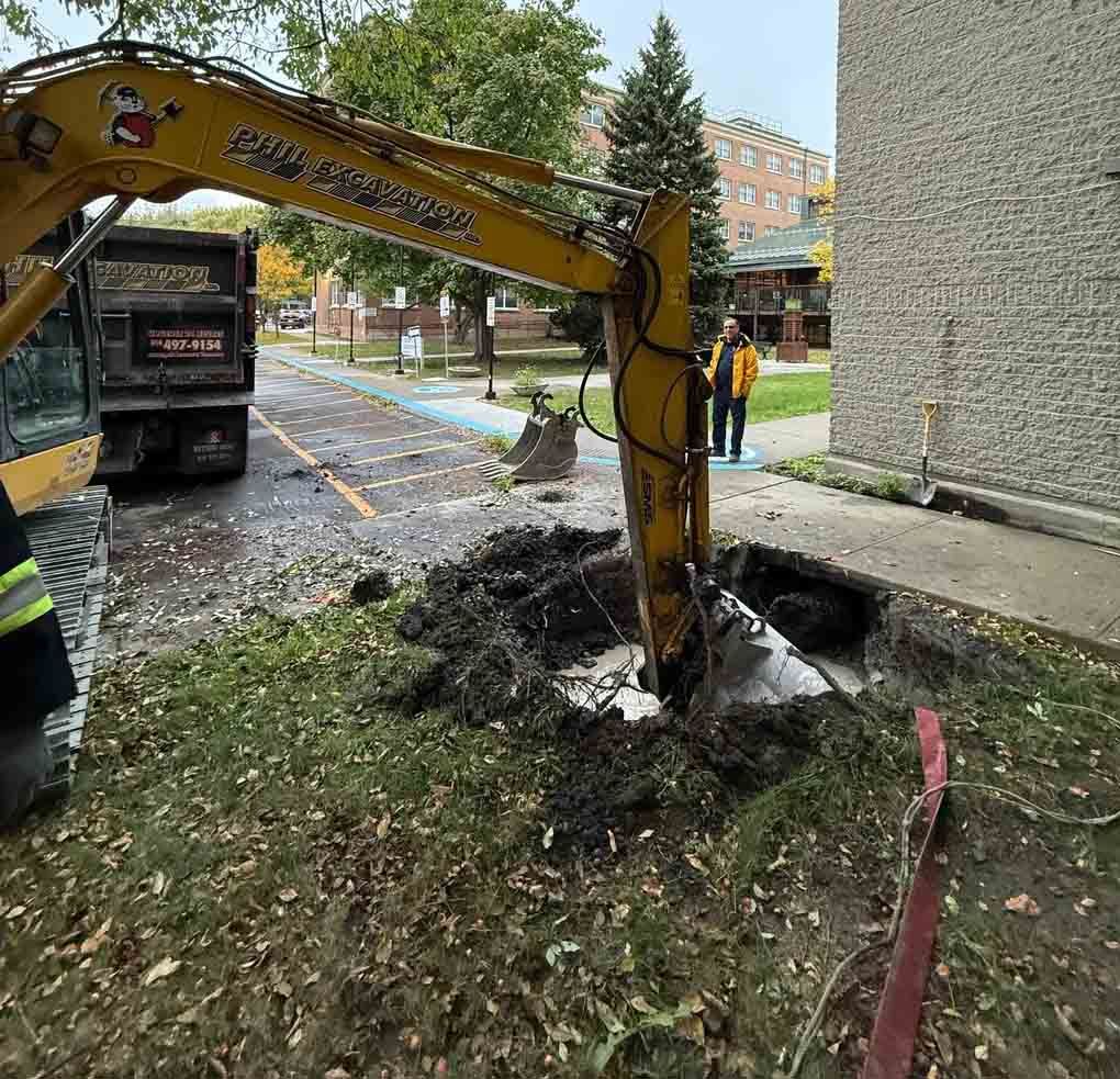 Construction site with an open trench, trench box, backhoe, and orange vehicle.