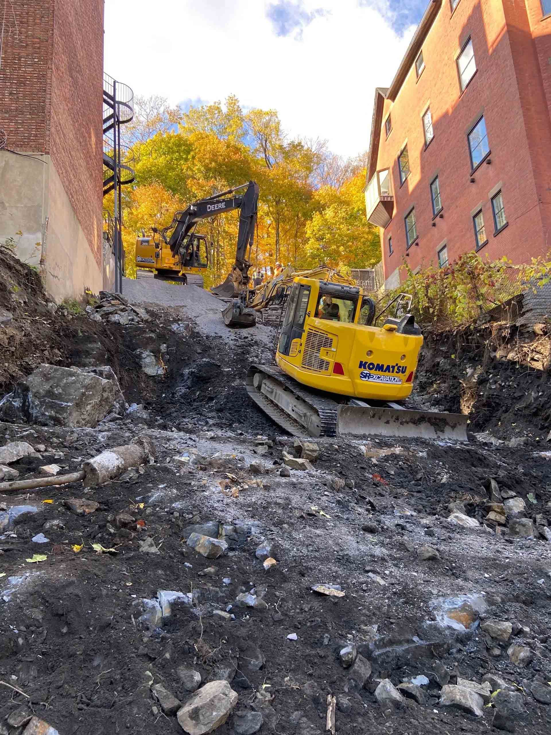 Construction site with two yellow excavators on a steep, rubble-covered street. Buildings on either side.