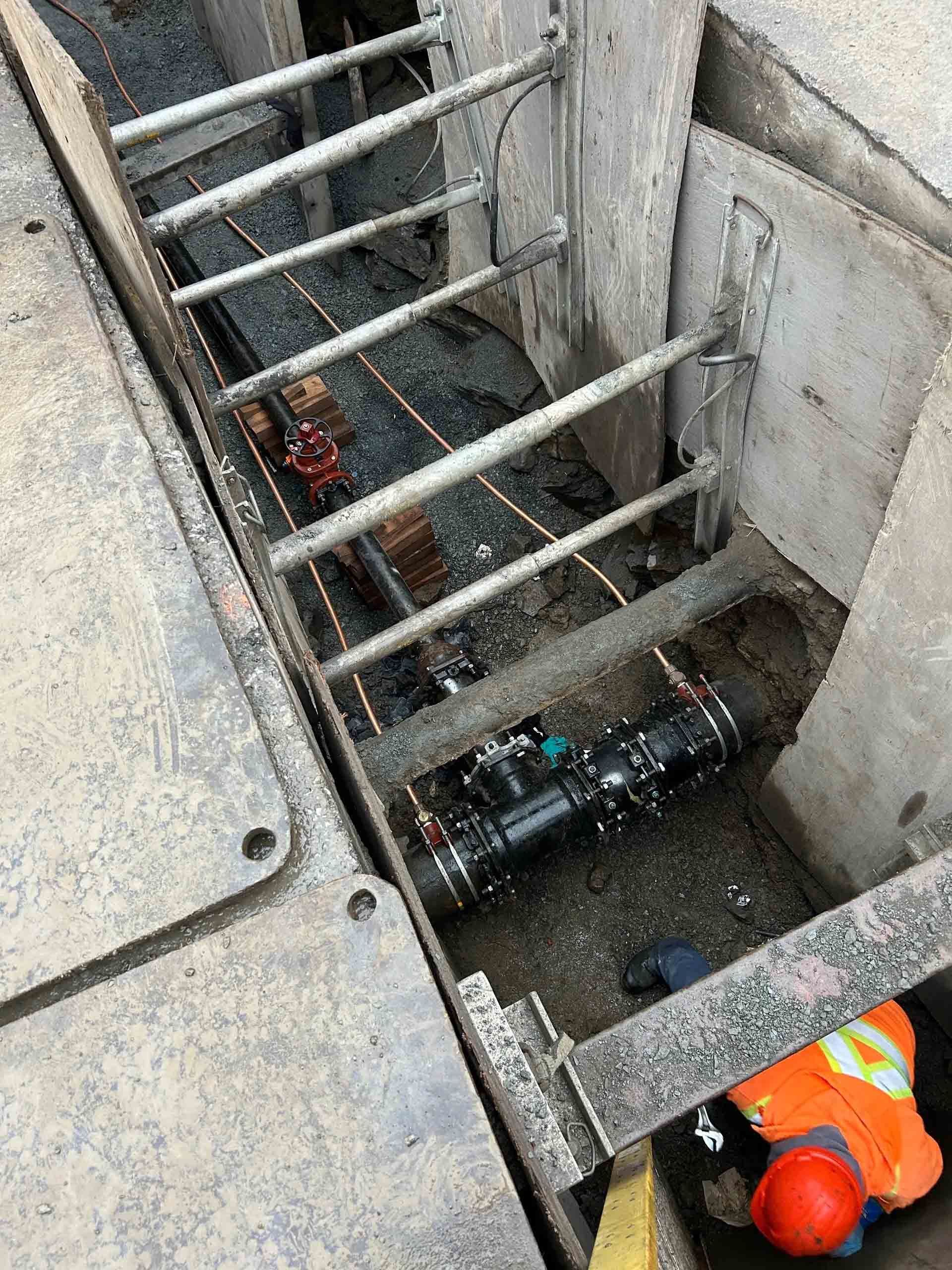 Chantier : tranchée avec étaiement, échelle et pelle mécanique. Un ouvrier travaille dans la tranchée. Un camion orange et un bâtiment sont visibles en arrière-plan.