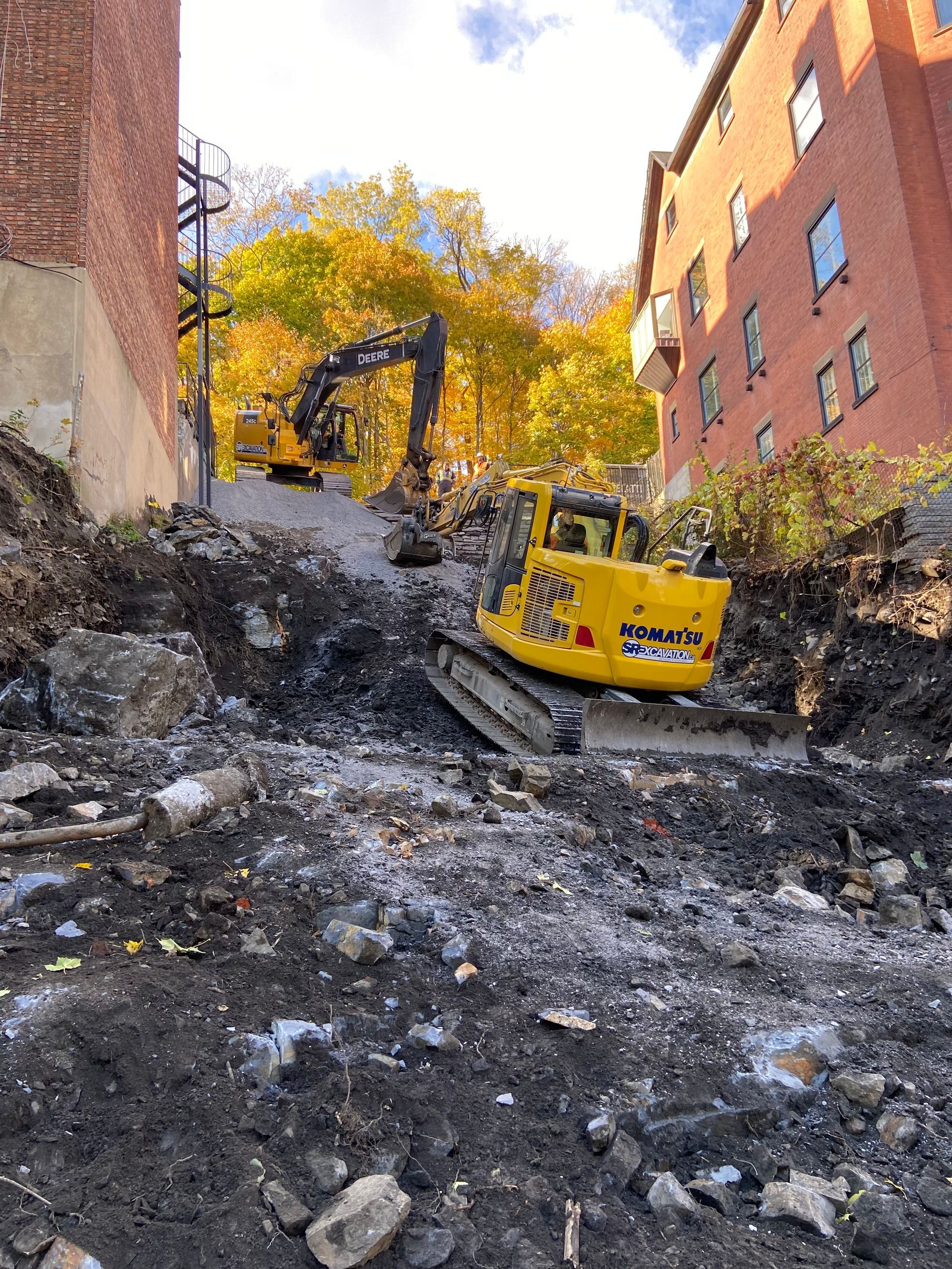 Construction site with two yellow excavators on a steep, rubble-covered street. Buildings on either side.