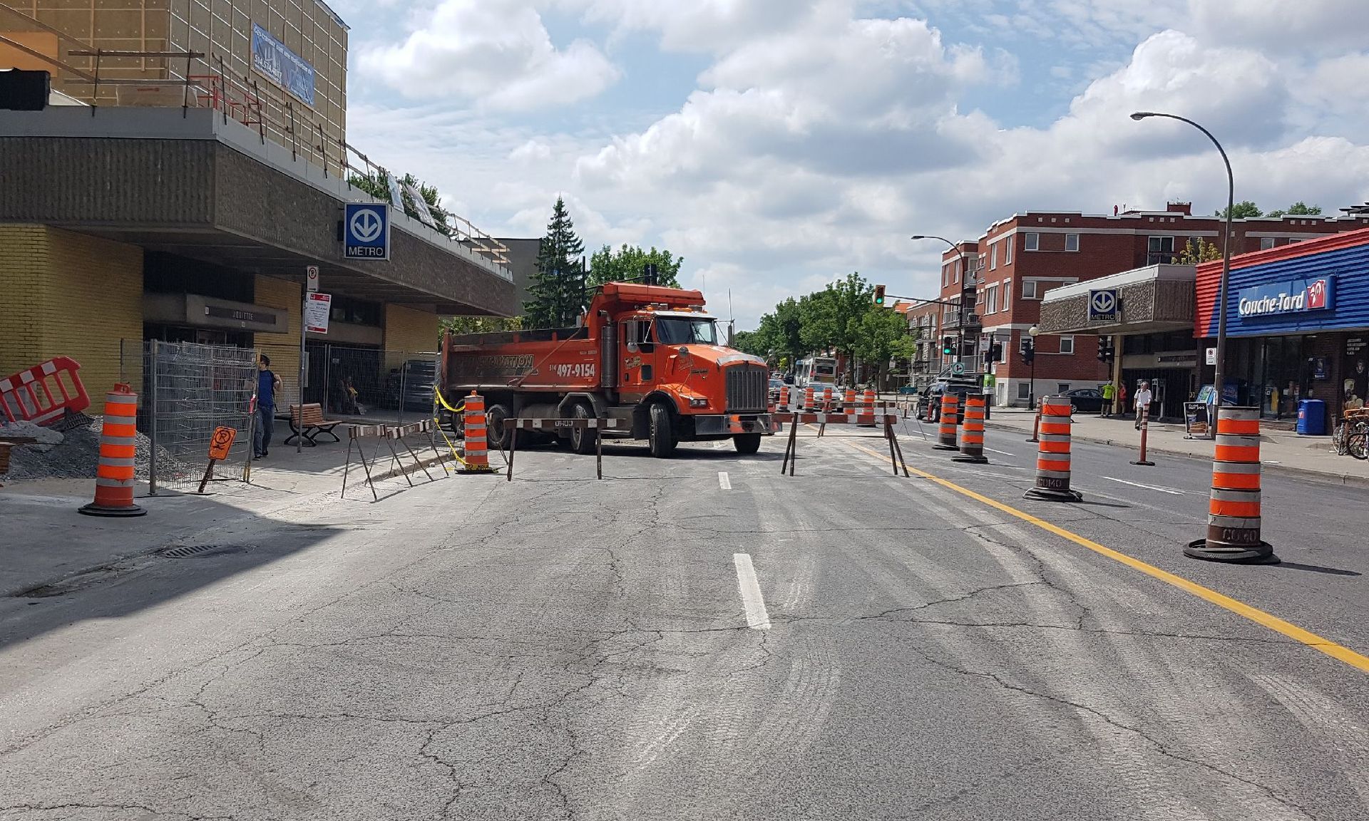 Scène de travaux routiers : camion-benne orange sur une rue partiellement fermée, pylônes orange, bâtiment en arrière-plan.