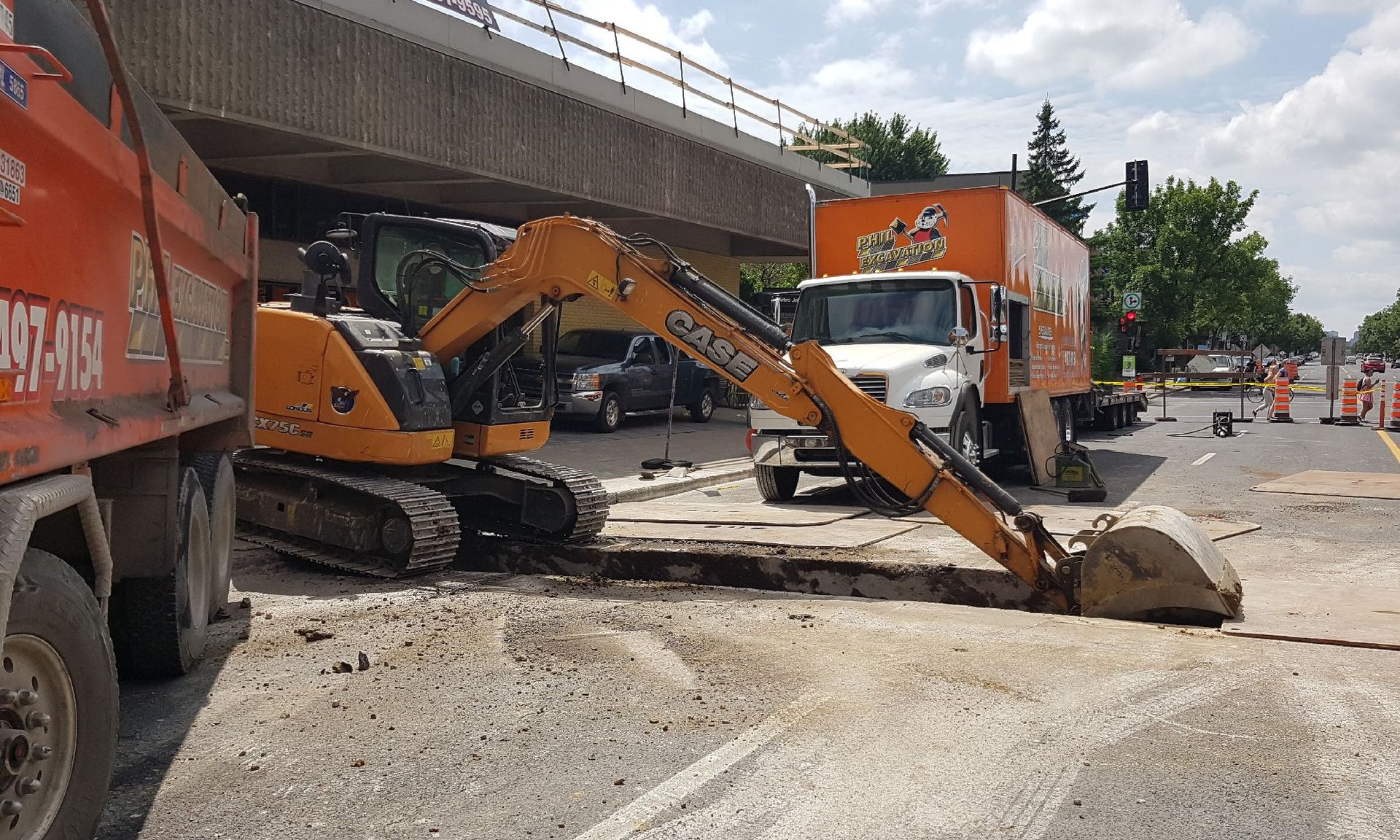 Excavator digging in road under overpass; trucks and construction zone setup.