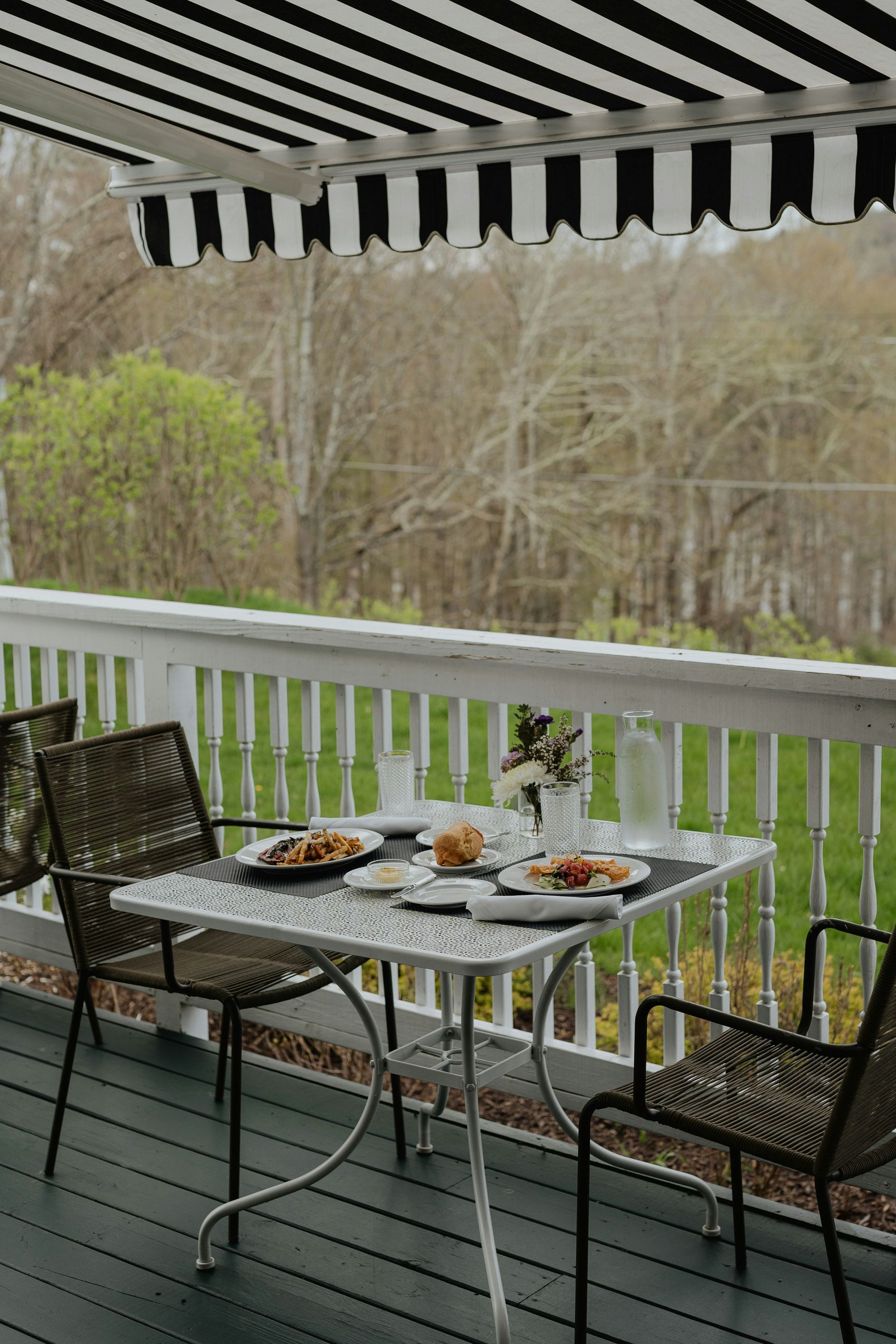 Patio table set for two with food, under a black and white awning, overlooking a wooded area.
