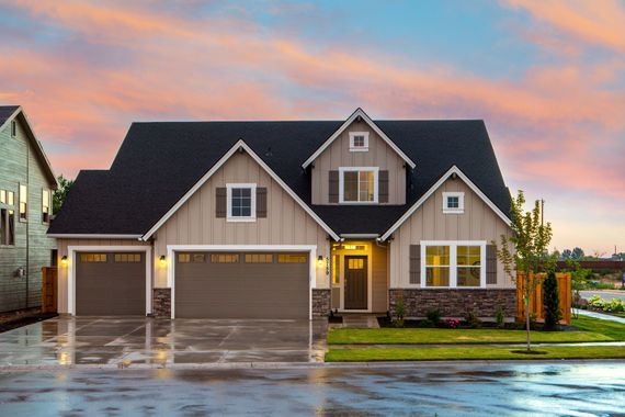 Tan house with black roof, three-car garage, and stone accents, set against a sunset sky.