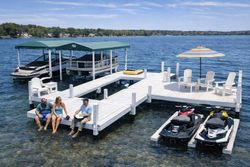 Three people sit on a white dock on a sunny lake, with boats, jet skis, and patio chairs nearby.