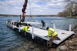 Workers in neon vests use a crane to install a white modular dock into a lake.