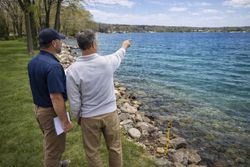 Two people stand on a grassy lakeshore, one pointing toward the water, with a survey marker visible on the rocky bank.