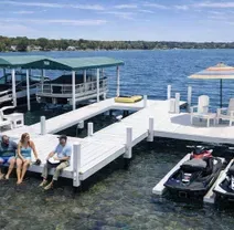 Three people sit on a white dock on a lake with two covered boats, two jet skis, and patio furniture under an umbrella.