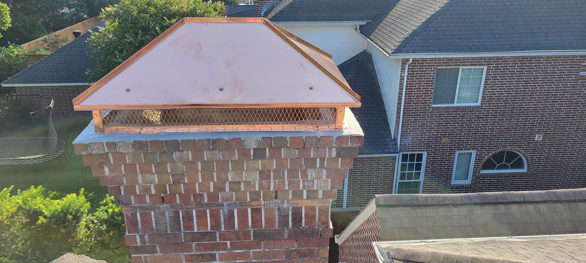 A copper chimney cap sits atop a brick chimney. A house with dark roof is in the background. Green trees.