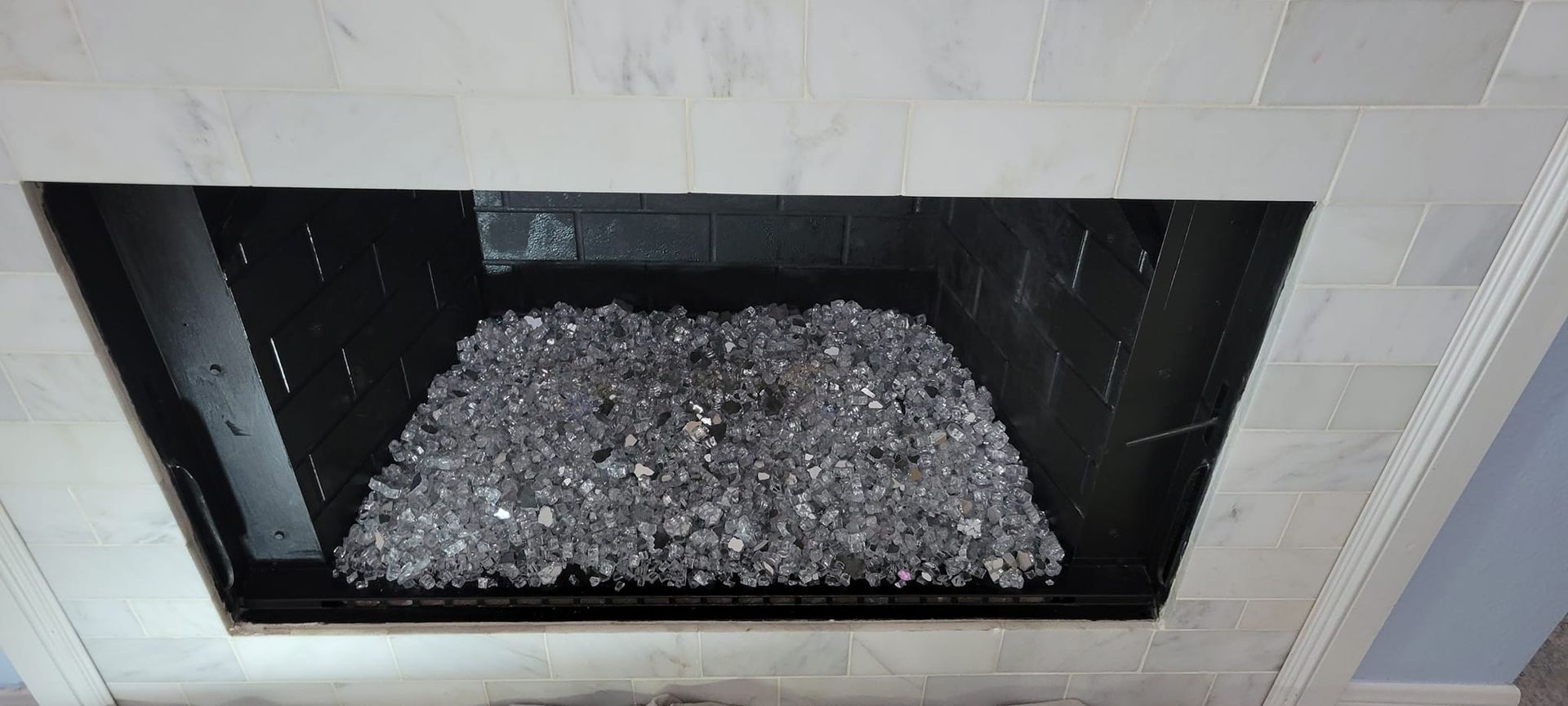 A fireplace with black interior and clear glass rocks. It is bordered by white stone tile.
