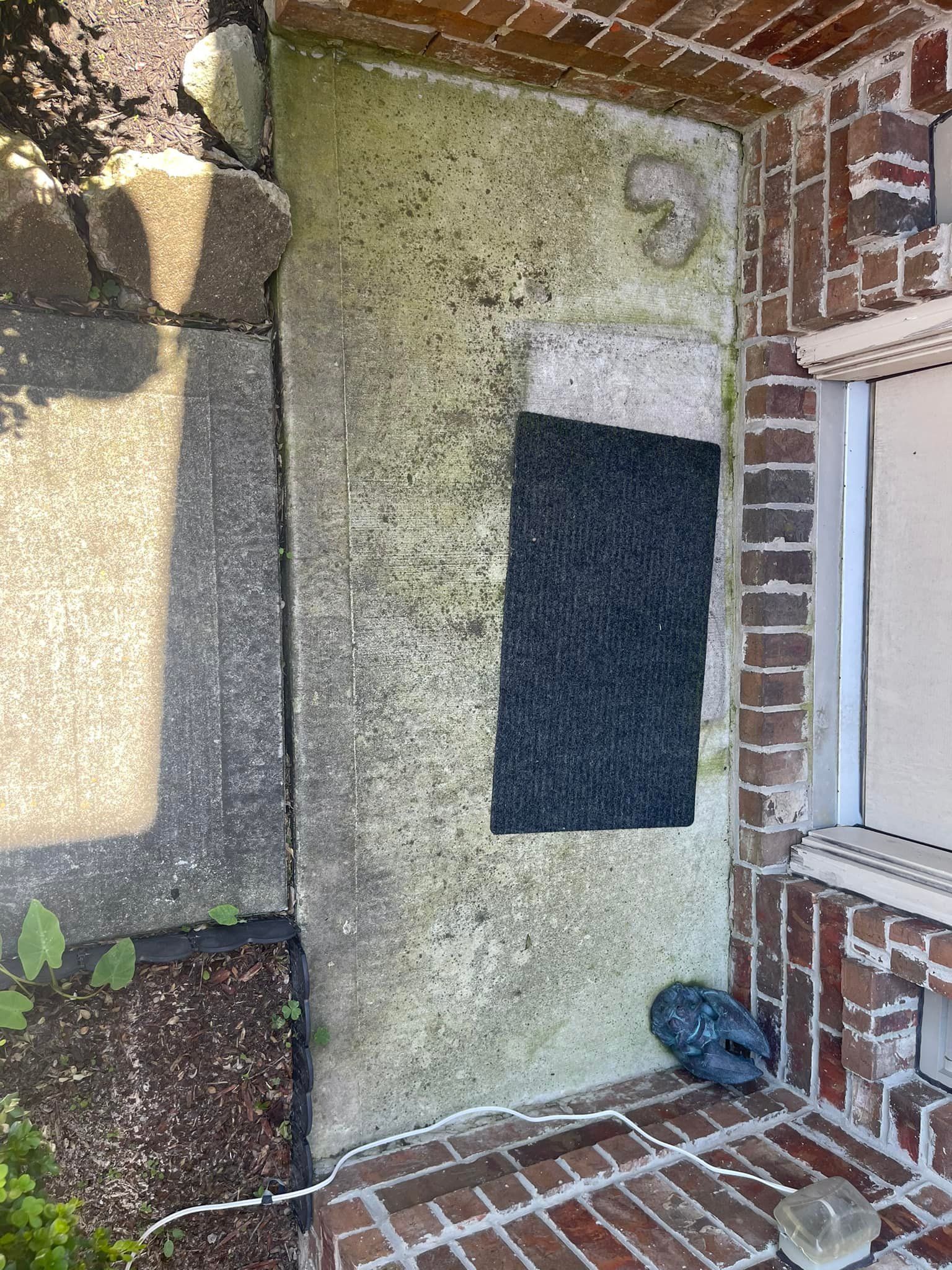 Weathered headstone with a black rectangular plaque, next to a brick wall.