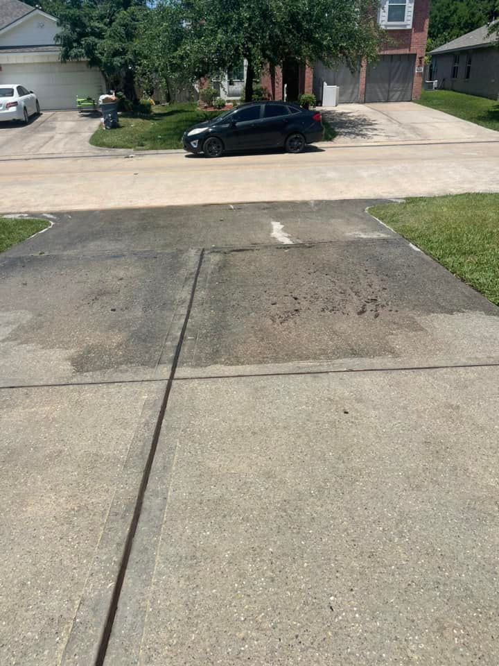 Concrete driveway with dark stains, black car parked, street, and houses in background.