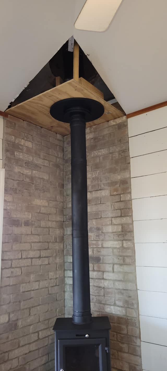Black stove with chimney pipe going through a wooden platform in a brick corner.