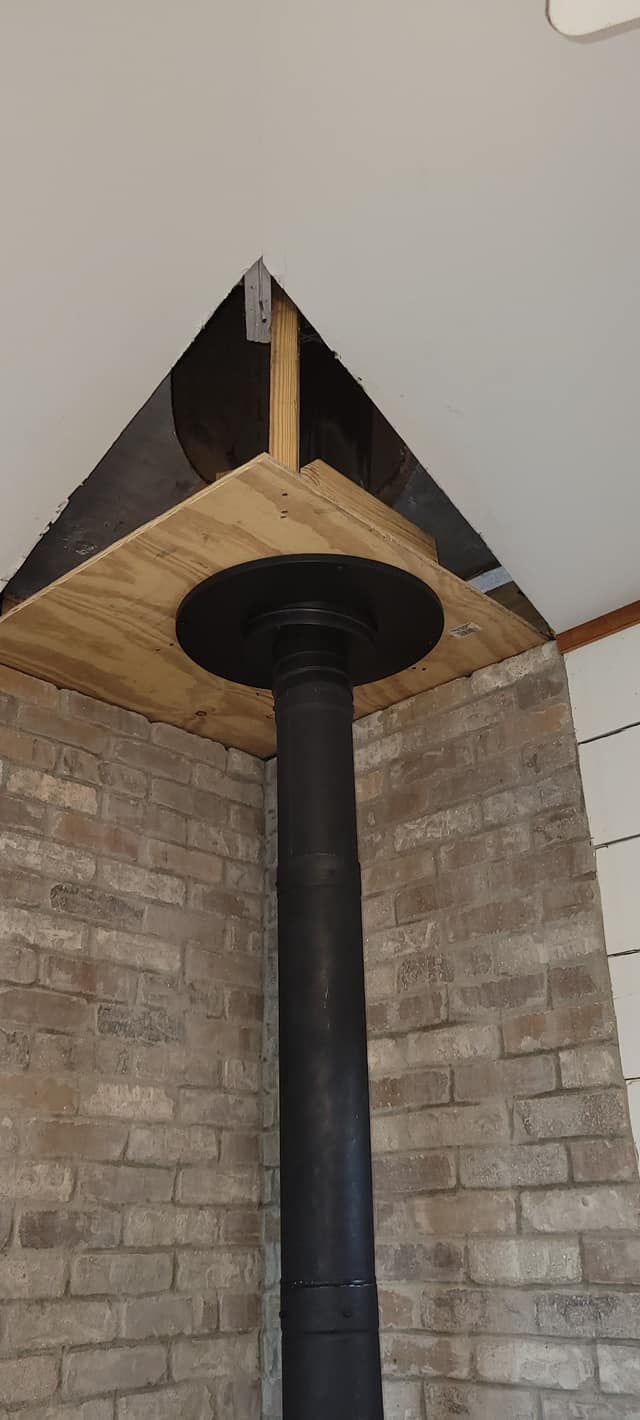A black chimney pipe in a brick corner, with a wood and black metal plate at ceiling.