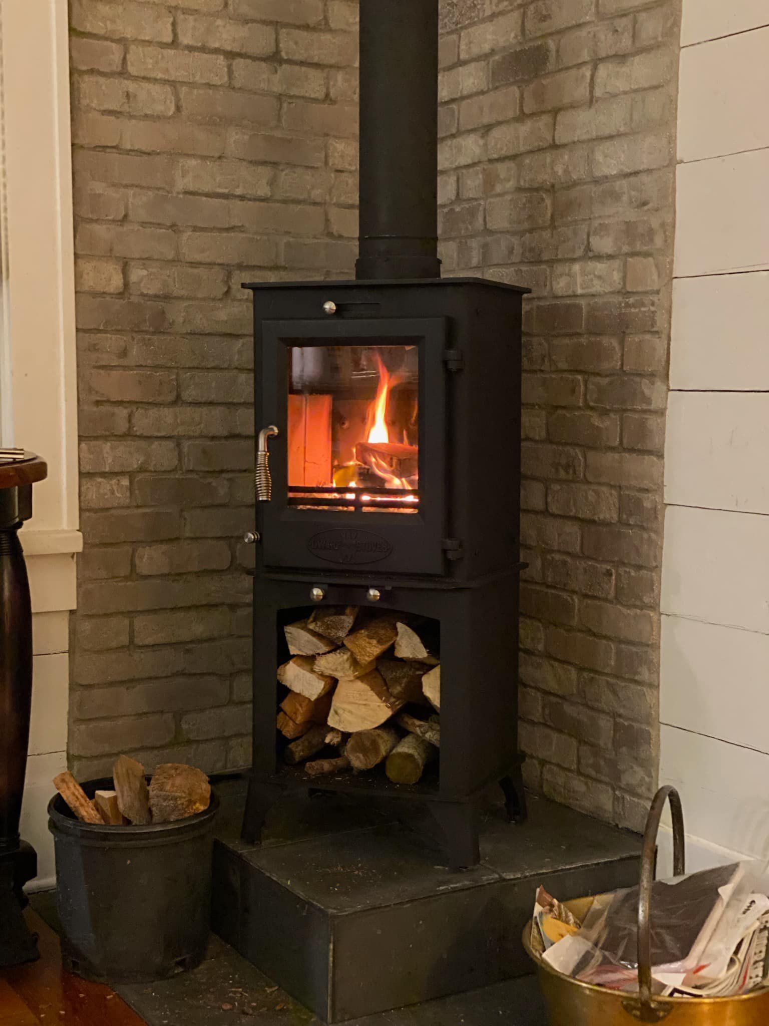Black wood-burning stove with lit fire, logs inside and below, set against a brick wall.