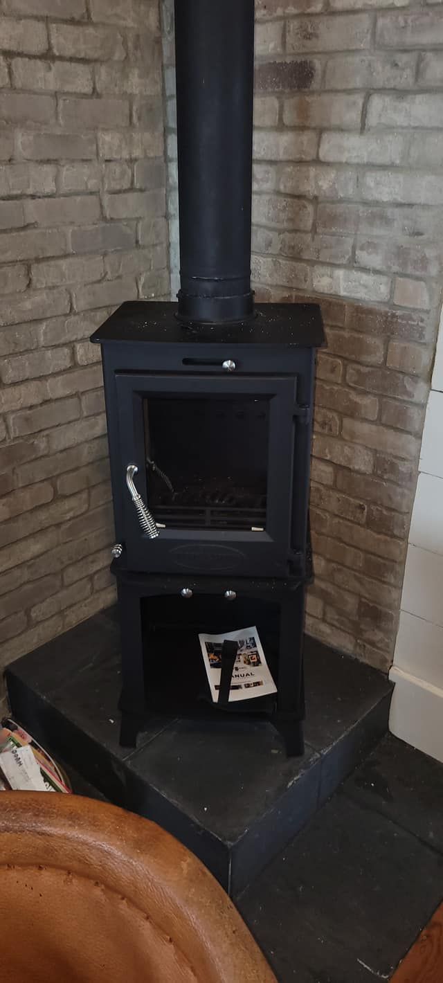 Black wood-burning stove with flue, placed in a corner against a brick wall. A magazine sits on the lower shelf.