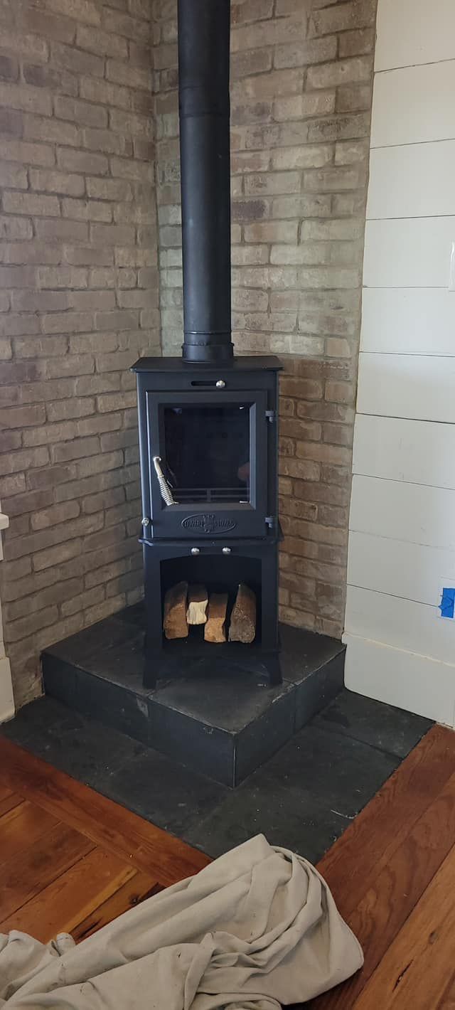 Black wood-burning stove with a flue pipe on a black stone hearth in the corner of a room with brick and white paneled walls.