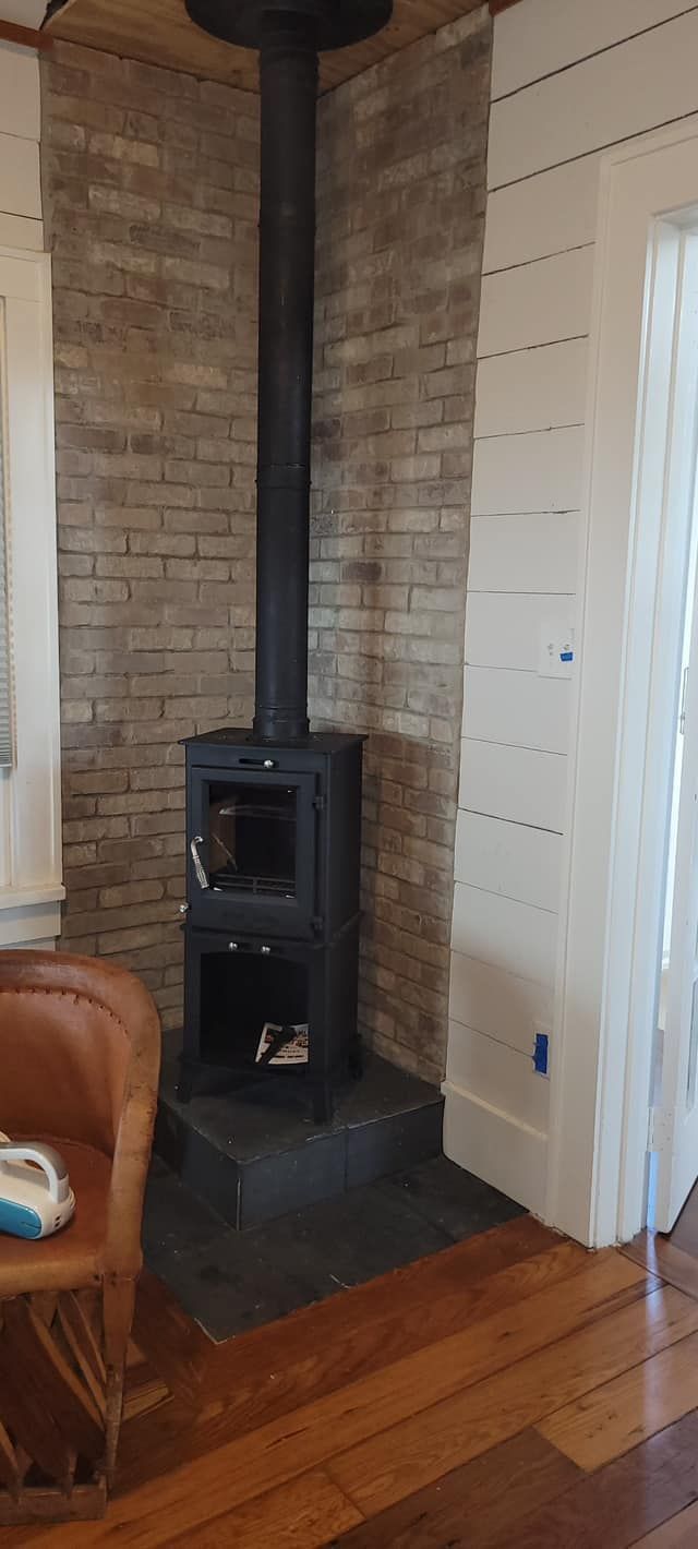 A black wood-burning stove with a chimney against a brick wall in a room with hardwood floors.