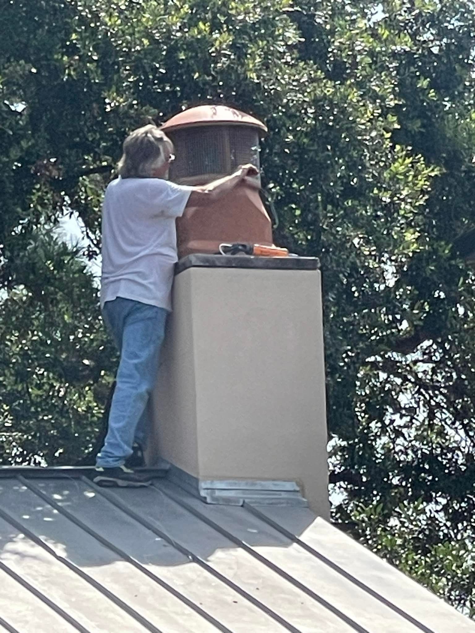 Man on rooftop, working on chimney cap. Gray roof, trees in background.