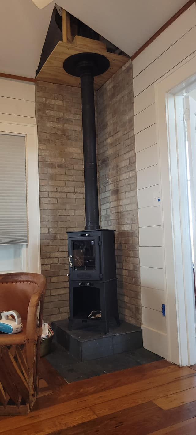 A wood-burning stove with a chimney against a brick wall. The floor is wooden.