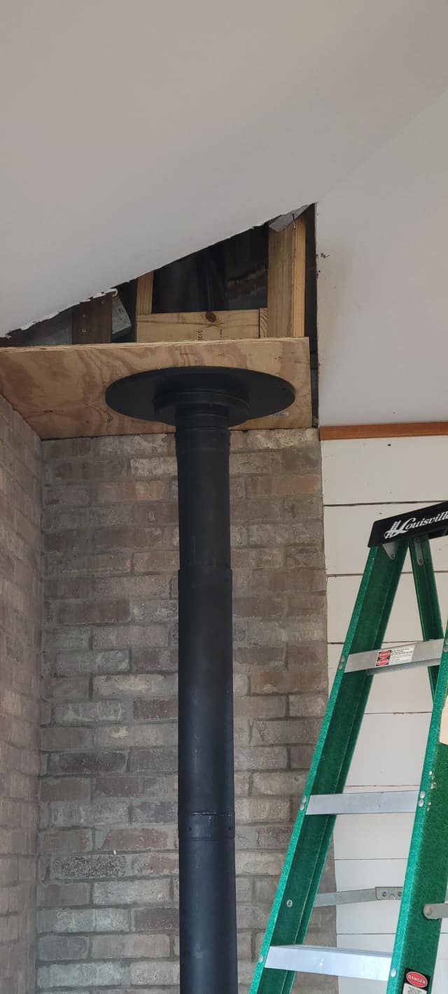 Black stove pipe through a ceiling, resting on brick wall; green ladder nearby.