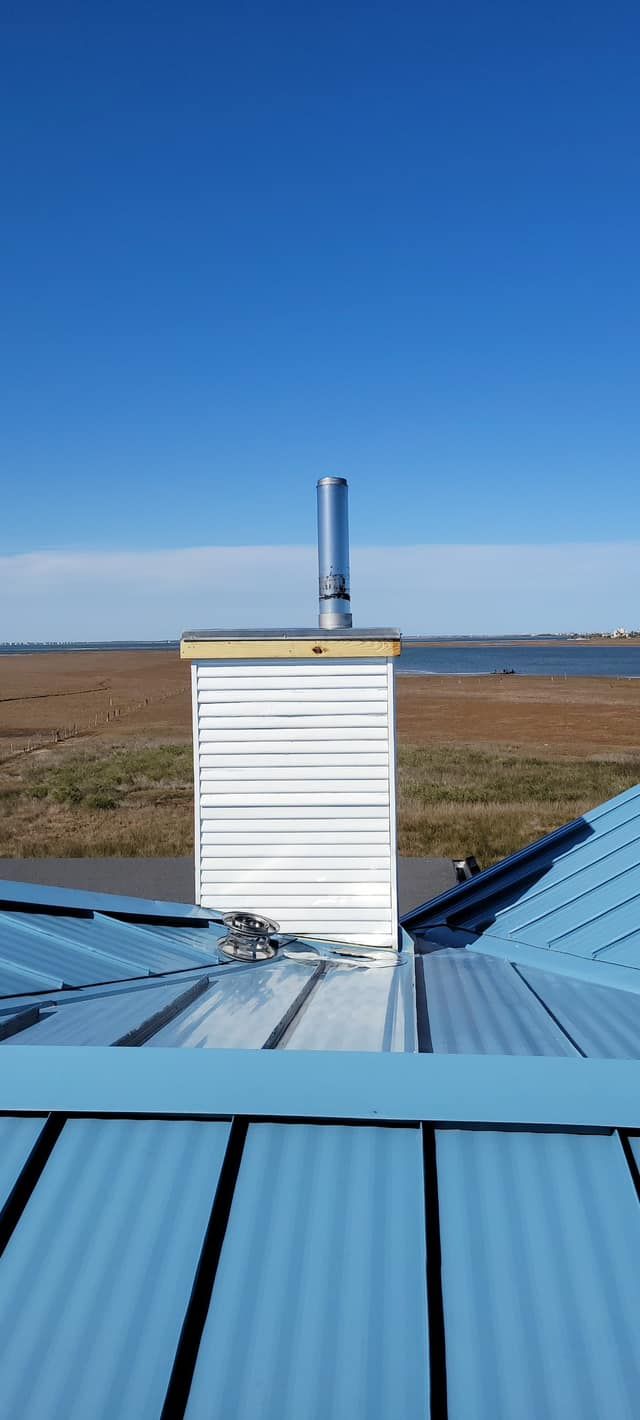 A metal chimney on a blue corrugated metal roof against a blue sky with land and water in the background.