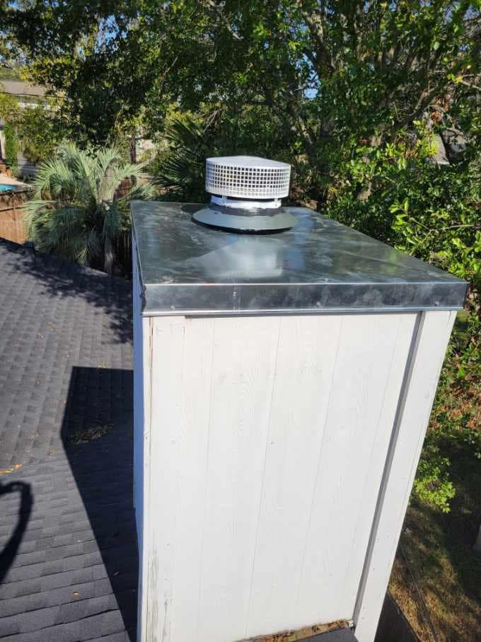 White chimney with metal cap and vent on a rooftop, with trees in the background.