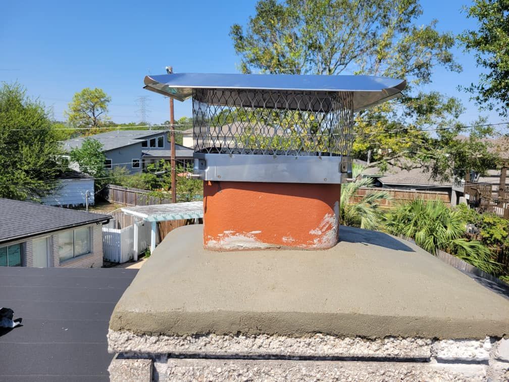Chimney with a metal mesh cap on a rooftop, with surrounding houses and trees.