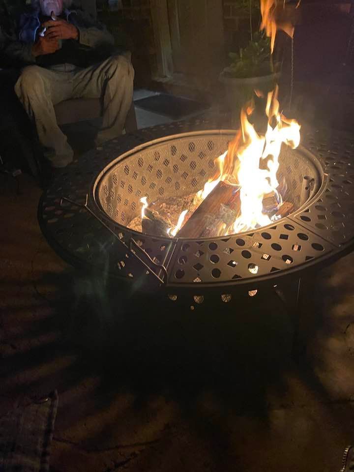 Fire pit with burning logs, casting shadows on patio. Person seated nearby.