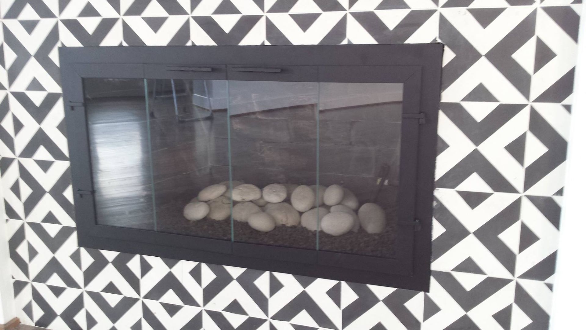Fireplace with a black metal frame, glass door, and white stones inside, set against a black & white geometric tile pattern.