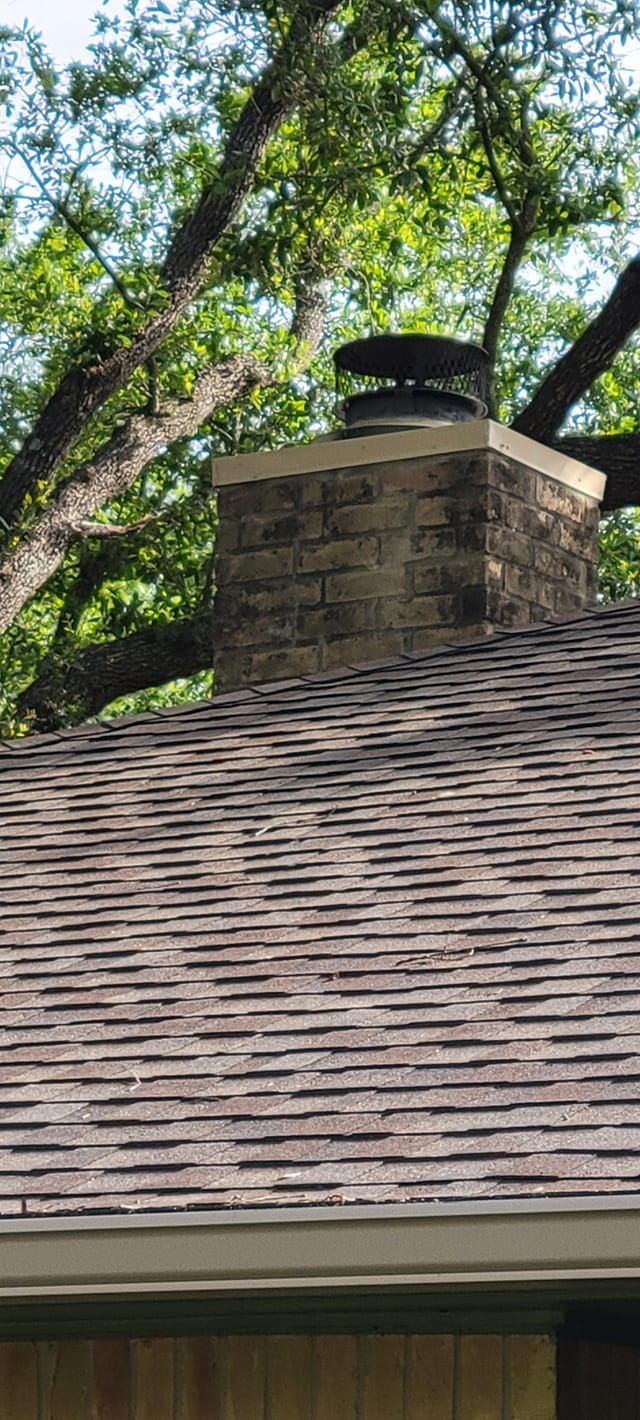 Chimney on a brown shingled roof with a tree in the background.