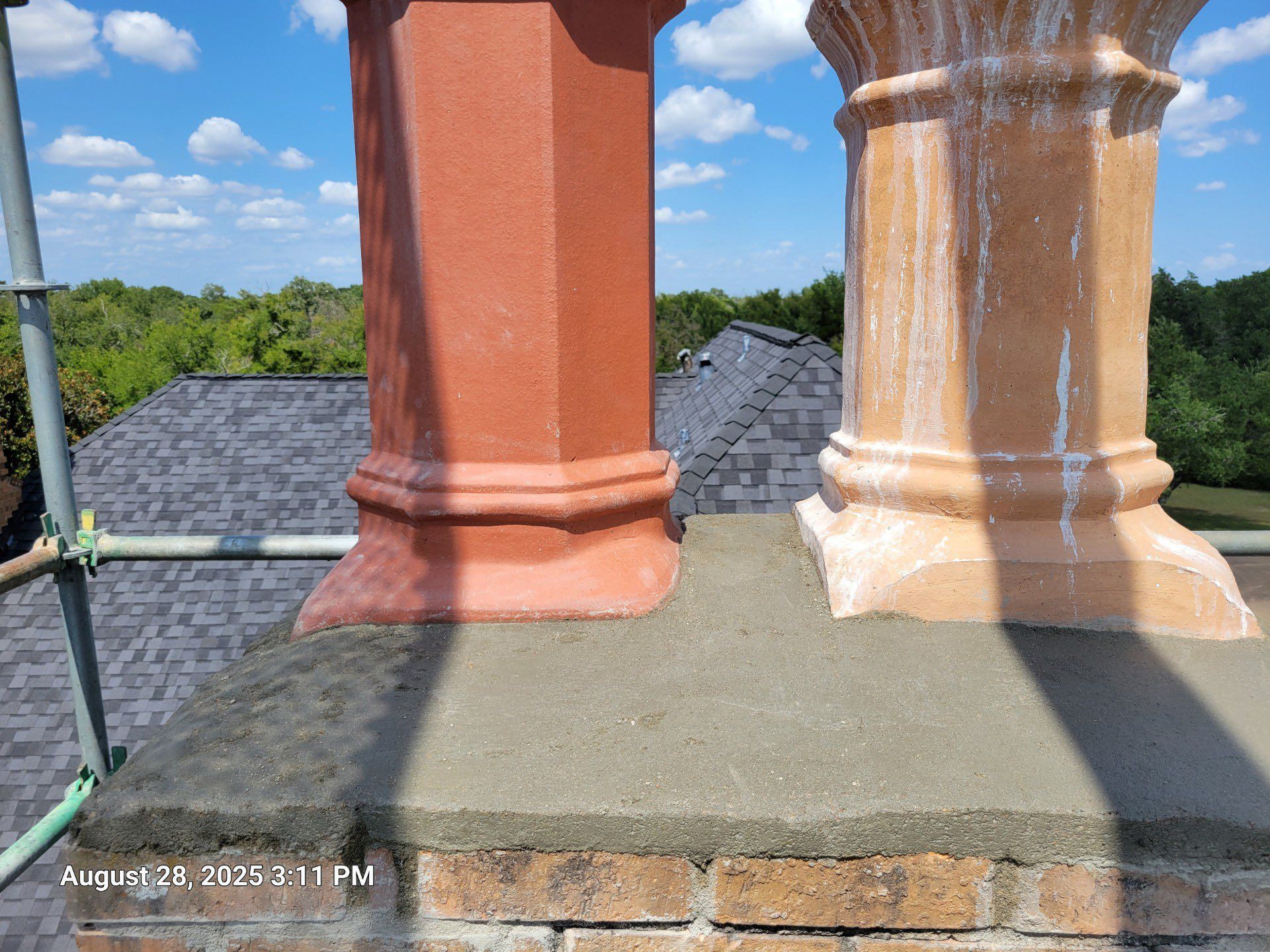 Two chimneys, one red, one tan, atop a brick base, against a blue sky with trees.