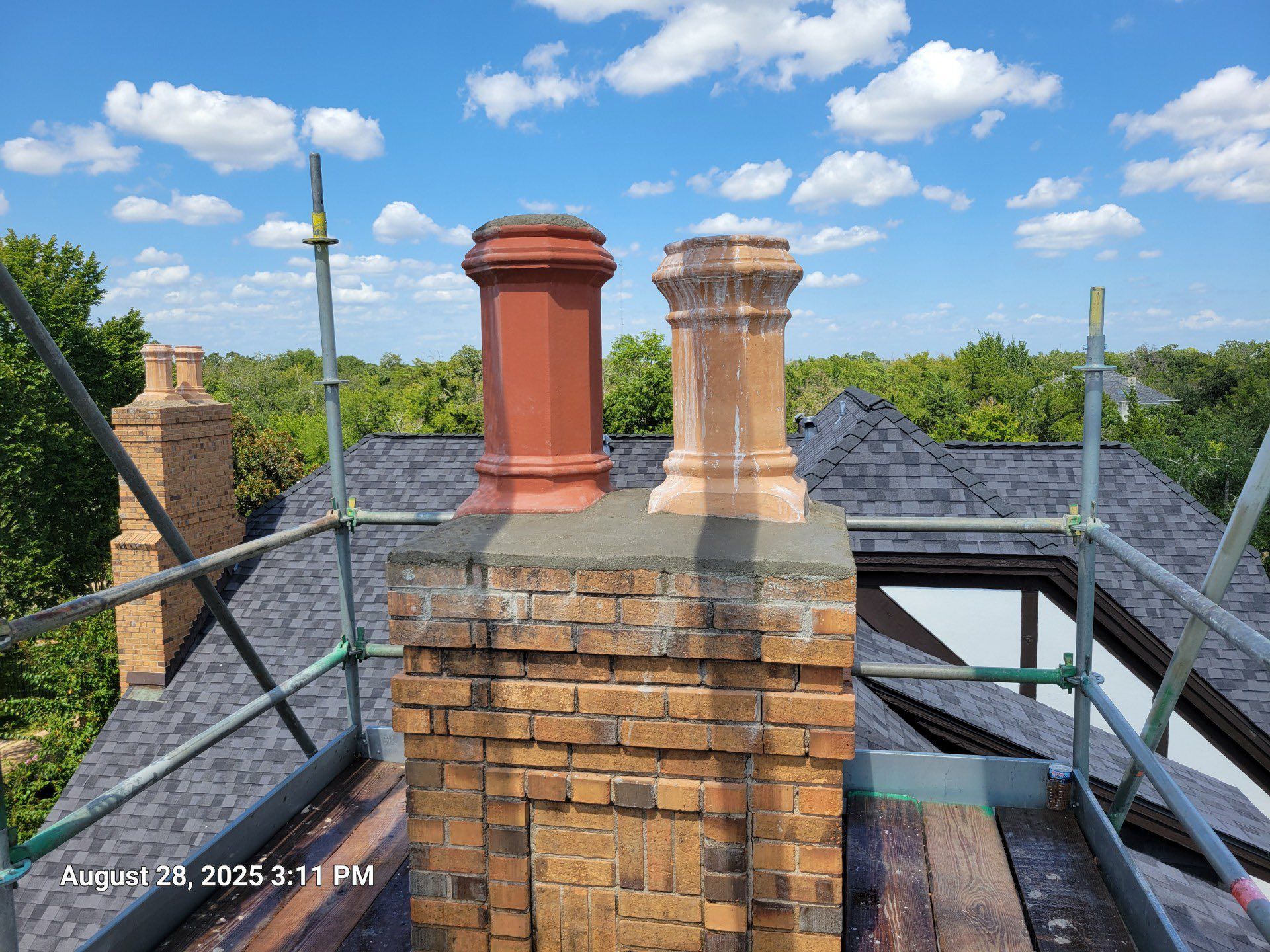 Two chimney pots on a brick chimney with scaffolding, set against a blue sky with clouds and a treeline.